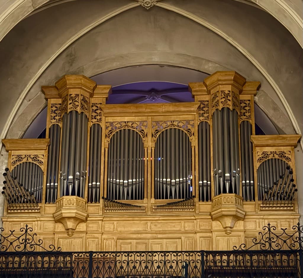 A historic pipe organ with carved wooden casework and polished metal pipes, framed by a stone arch inside a Catholic church.